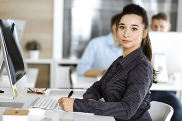 Business woman using computer at workplace in modern office. Brunette secretary or female lawyer looking at the camera and happy smiling. Working for pleasure and success