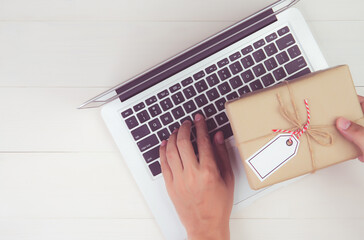 Hand of man holding gift box and tag having laptop computer on wooden table, present giving for Christmas day or birthday, season and celebrate, top view, flat lay, online shopping concept.