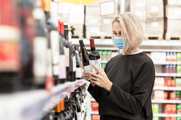 Young blonde woman in a medical mask chooses a bottle of wine in a supermarket. Coronavirus pandemic.