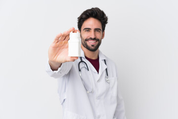 Young handsome man with beard over isolated white background wearing a doctor gown and holding pills