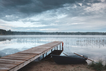Morning landscape. The shore of the lake with a wooden pier and tied the boat at dawn. The water reflects the sky. A light fog is spreading over the water. As a concept of outdoor recreation.