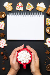 Hands hold red ceramic cup with cocoa and marshmallows on background of christmas cookies with copy space