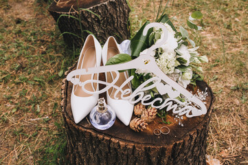 Wedding accessories and details of the bride close-up: white hanger, leather shoes, bouquet, rings, cones, hairpin, perfume. Female set on a wooden stump, photography, concept.