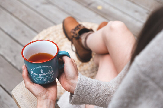 The Girl Is Sitting On The Wooden Terrace Under A Blanket And Drinking Hot Tea. Her Feet Are In Brown Boots With The Laces Untied. In The Hands Of A Mug With A Motivational Inscription.