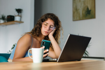 Bored young woman working at home with laptop and using smartphone.