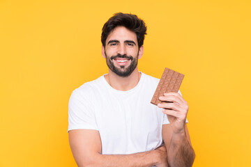 Young handsome man with beard over isolated yellow background taking a chocolate tablet and happy