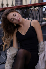 attractive slim young brunette in fashionable trench coat and black dress sitting on a bench. autumn, yellow leaves.