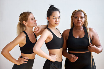 Fototapeta na wymiar three young diverse multi ethnic female models of different race, hair colour and body size posing at camera, friendly females in black sportswear. tolerance and natural beauty concept
