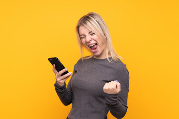 Young blonde woman over isolated yellow background with phone in victory position