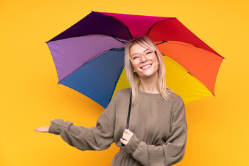 Young blonde woman holding an umbrella over isolated yellow wall holding copyspace imaginary on the palm