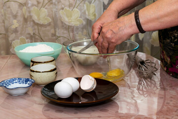 An elderly woman breaks an egg with a knife into a glass bowl for making pancake batter.Ingredients on the table-milk, salt, sugar, wheat flour, butter.Selective focus.