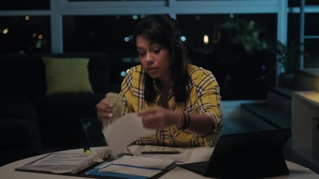 Latin American woman doing home budget and counting money, girl checking cash to pay utility bills late at night. Young hispanic lady reviewing invoices, doing family business plan and tax return