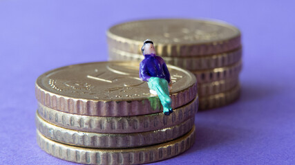 Miniature figurine of man sitting on stack of euro cent coins with another pile of coins in the back in front of violet background
