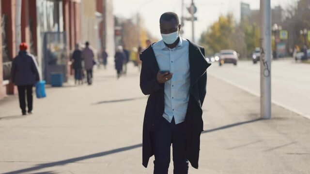 Portrait of African American business man in mask, smartphone in hands. City walk down the street during the coronavirus pandemic.