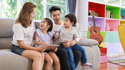Happy Asian family teaching children son and daughter how to use tablet while sitting on grey sofa in living room with smiling faces