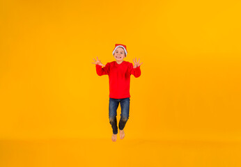 Obraz premium happy little boy in red Christmas hat jumps on yellow background with space for text