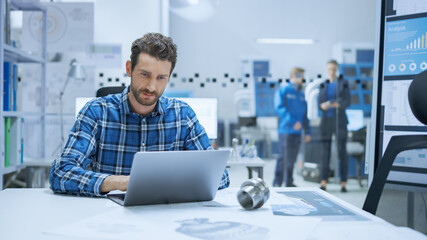 
Modern Industrial Factory: Industrial Engineer Sitting at His Desk, Working on Laptop Computer, Analyzing Mechanism and Blueprints. in Background Functional Manufactory with CNC Machinery