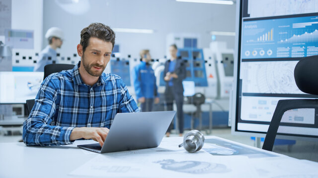 Modern Industrial Factory: Industrial Engineer Sitting at His Desk, Working on Laptop Computer, Analyzing Mechanism and Blueprints. in Background Functional Manufactory with CNC Machinery
