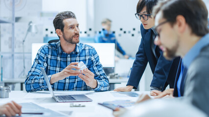 Modern Factory Office Meeting Room: Multi-Ethnic and Diverse Team of Engineers, Managers and Investors Talking Sitting at Conference Table, Analyzing Blueprints, Showing Information on Laptop Computer