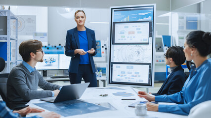Modern Industrial Factory Meeting: Confident Female Engineer Uses Interactive Whiteboard, Makes Report to a Group of Engineers, Managers Talks and Shows Concept of Sustainable Eco-Friendly Engine
