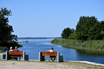 View of a coast of a vast river or lake merging with the Polish sea with two grass and tree covered banks or coasts to be seen on both sides and some tourists resting on wooden benches on the pier