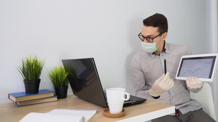 Young Caucasian male wear protective disposable face mask, white gloves sits at table, uses laptop, tablet, prepares presentation about growth chart on stock exchange in lock down period. Remote work. - Powered by Adobe