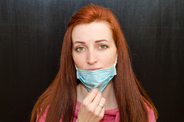 Young caucasian redhaired girl with green eyes, in medical mask, pink sweater on black background,...