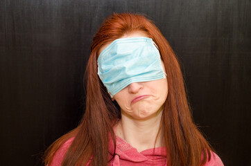 Young caucasian redhaired girl with green eyes, in wrong used medical mask, on black background,...