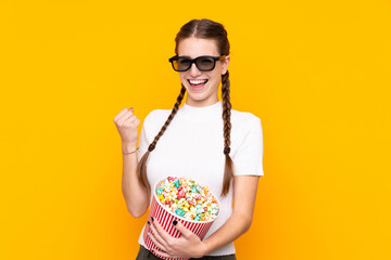 Obraz premium Young woman over isolated yellow background with 3d glasses and holding a big bucket of popcorns