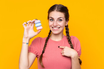 Young woman over isolated yellow background holding colorful French macarons with surprise expression