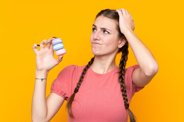 Young woman over isolated yellow background holding colorful French macarons with confuse expression