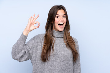 Teenager Brazilian girl over isolated blue background surprised and showing ok sign