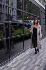 Fototapeta premium beautiful slim young brunette in fashionable trench coat and black dress walking through the street. autumn, yellow leaves