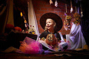 Obraz na płótnie Canvas Little cute blonde girl looking as witch in special dress and hat in room decorated for Halloween. Witchcraft and wizardry in carnival. Halloween style photo shoot.