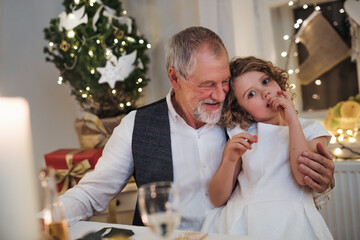 Senior grandfather with small granddaughter indoors at Christmas, sitting at table.