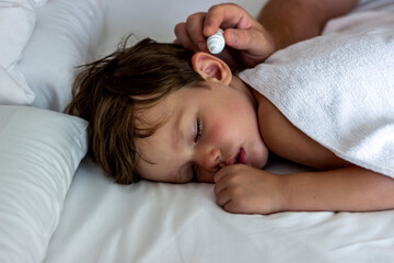 Obraz na płótnie Canvas Father dribble medication into boy ear.Photo of a dad dripping ear drops to teenager son while sleeping in the comfortable bed at home.Ear drops.Close up man drips medicine into the ear of a sick boy.