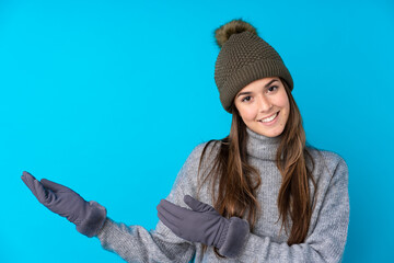 Teenager girl with winter hat over isolated blue background extending hands to the side for inviting to come