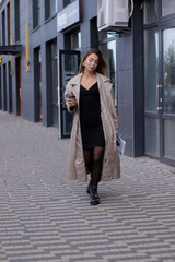 Obraz premium beautiful slim young brunette in fashionable trench coat and black dress walking through the street. autumn, yellow leaves