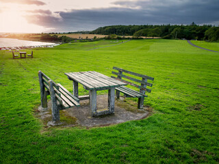 Old style table and benches in a park in focus, Car park and ocean out of focus in the background. Nobody, cloudy sky with sun flare.