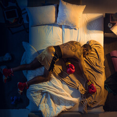 Kicking. Top view of young professional boxer, fighter sleeping at his bedroom in sportwear with gloves. Loving his sport even more than comfort, playing match even if resting. Action, motion, humor.