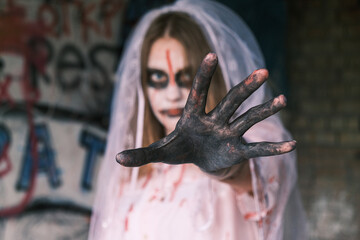 Horror, nightmare and Halloween concept. Dead bride. Young woman in zombie makeup with bride's veil on her head looks into camera reach out her hand forward