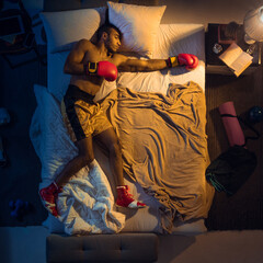 Punching. Top view of young professional boxer, fighter sleeping at his bedroom in sportwear with gloves. Loving his sport even more than comfort, playing match even if resting. Action, motion, humor.