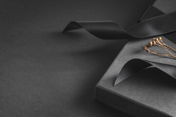 Black gift box on a black dark background, decorated with a textured bow and dry branch. Present...