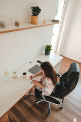 View into a beautiful light home office during working week. Woman in pajamas is sitting on a chair and she is making notes. She is keeping social distance since he covid-19 pandemic. 