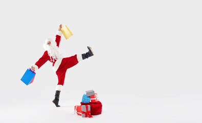 Joyful Santa walks widely with a bag of gifts and shopping bags on his back on a white background. New Year and Christmas holidays and sales, place for text, banner.