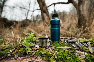 Fototapeta premium Camping vacuum thermos with a mug in the forest. Autumn forest, green moss on the tree