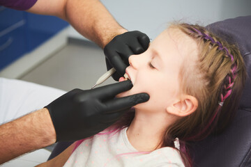 Fototapeta premium Portrait of little girl has a dental examination while sitting at dentist office. Male dentist is checking the young patient's mouth.