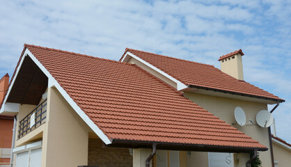 Fototapeta premium A close-up on a stucco house with a red metal tiled double gabled roof, a chimney, fascia boards, rain gutters, two satellite dish antennas and an attic open balcony against blue sky.