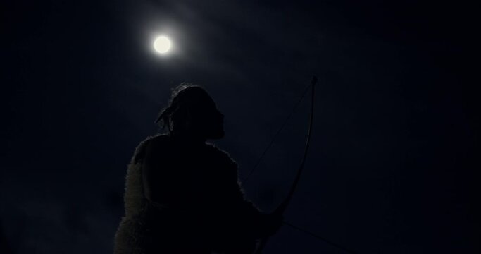 Primitive man hunting at night in full moon. From below side view of primeval male with bow and arrows hunting at night in nature on background of dark sky with full moon