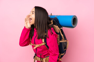 Young mountaineer girl with a big backpack over isolated pink background shouting with mouth wide open to the lateral